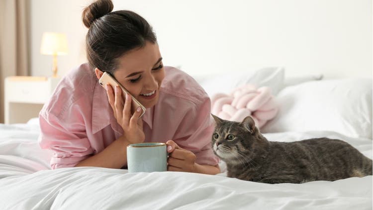 A cat owner talks on the phone while her pet relaxes beside her.