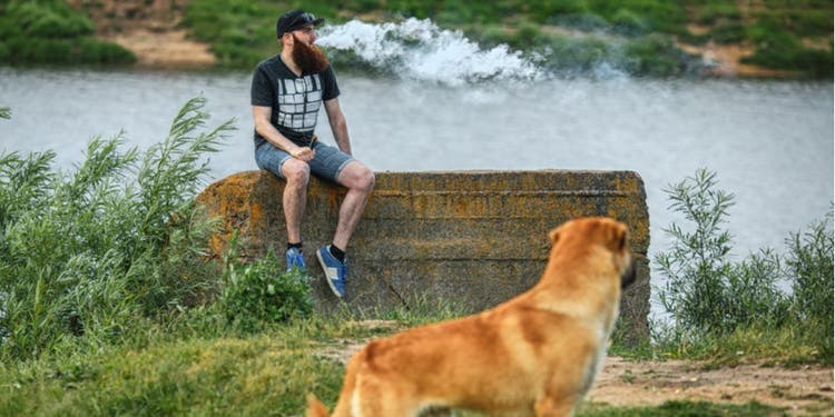 A man exhales a vape cloud while a dog watches from a distance.