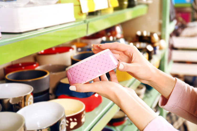 A woman examines a pink, ceramic dog bowl. A shelf in the background contains several more of various sizes and colors.