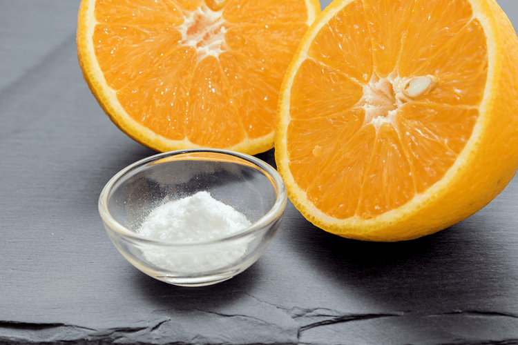 A sliced orange and a small bowl of solid vitamin C (ascorbic acid) on a black, stone table